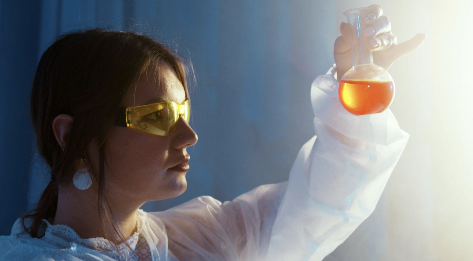 A young female scientist examining a flask with orange liquid in a laboratory setting. She wears protective golden goggles and a white lab coat, emphasizing safety in her work environment. 